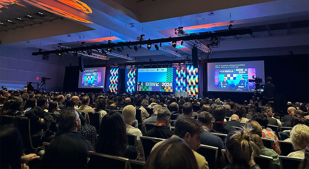 Großer Konferenzsaal beim SXSW 2024, gefüllt mit vielen Zuschauerinnen und Zuschauern. Auf der Bühne sind bunte Bildschirme mit der Aufschrift ‚SXSW 2024‘ zu sehen, daneben Leinwände mit einer Podiumsdiskussion und Kameraleuten im Vordergrund.
