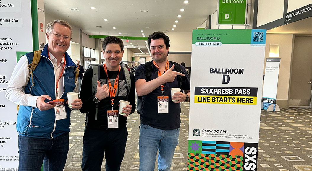 Brand Trust Gruppe von drei Personen steht vor einem Schild, das den Weg zum ‚Ballroom D‘ beim SXSW zeigt. Das Schild weist auf den Beginn der ‚SXExpress Pass‘-Warteschlange hin. Alle drei Personen halten Becher und tragen SXSW-Ausweise um den Hals. Im Hintergrund sind weitere Personen und ein Gang des Austin Convention Centers sichtbar.
