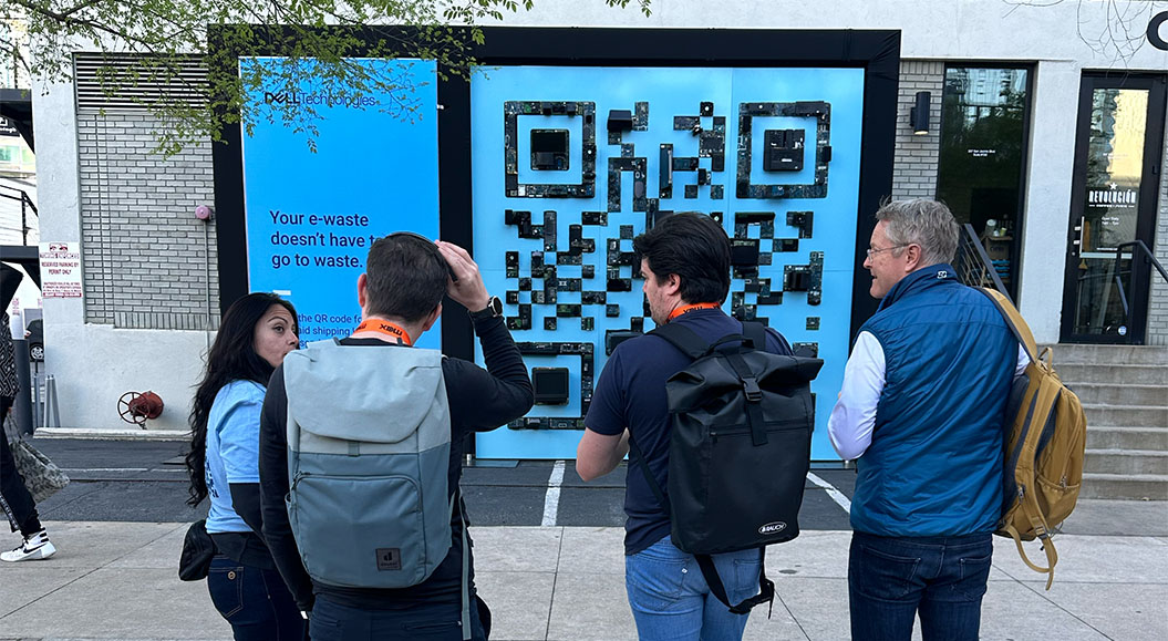 Eine Gruppe von vier Personen, die vor einer großen Wand aus elektronischen Bauteilen stehen, die ein QR-Code-Muster bilden. Auf der Wand ist der Text "Your e-waste doesn't have to go to waste" zu lesen. Es handelt sich um eine innovative Art der Recyclingwerbung, die das Thema E-Waste (elektronischer Abfall) aufgreift. Die Personen scheinen den QR-Code auf der Wand zu scannen oder zu betrachten. 