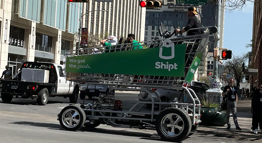 Ein großer Einkaufswagen von Shipt, der auf einem Fahrzeug montiert ist und in der Stadt auf der Straße fährt. Der Einkaufswagen hat eine grüne Werbefläche mit dem Text „We got the goods“. Drei Personen sitzen im Einkaufswagen, während zwei weitere Personen auf der Straße stehen und mit der Kamera fotografieren.