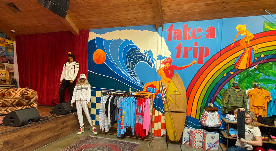 Das Bild zeigt einen lebendigen, modernen Laden mit einer bunten Wandmalerei im Hintergrund. Die Wand zeigt eine stilisierte Welle, die in leuchtenden Farben dargestellt ist, mit einer Person, die auf einem Surfbrett reitet, sowie einem Regenbogen und tropischen Pflanzen. Vor der Wand sind Kleiderständer mit bunter Mode, die aus T-Shirts, Hoodies und weiteren Kleidungsstücken bestehen. Auf der linken Seite des Bildes befinden sich zwei Schaufensterpuppen, die sportliche Kleidung tragen, und auf der rechten Seite ist eine Person, die ein Kleidungsstück hält. Das Gesamtbild vermittelt eine fröhliche, entspannte Atmosphäre mit einem Retro-Surfer-Thema.