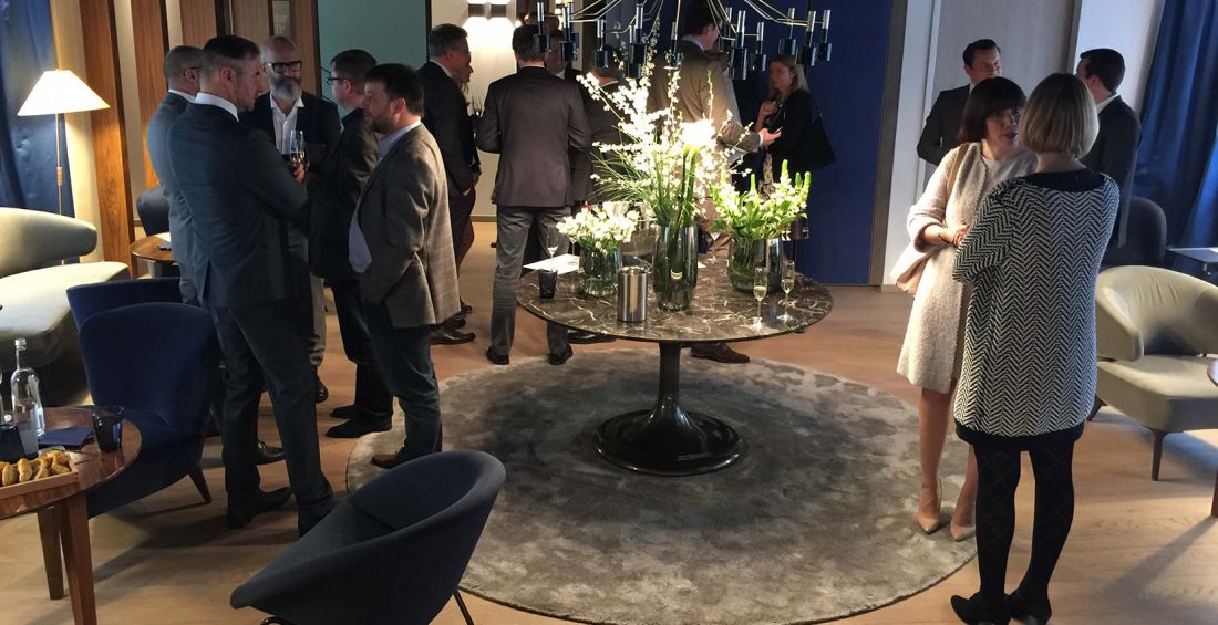 Networking im BrandTrist Nürnberger Büro: Eine Gruppe von Menschen steht in einem modern gestalteten Raum bei einem informellen Empfang zusammen. Mehrere Personen unterhalten sich im Stehen, einige mit Getränken in der Hand. In der Mitte des Raums steht ein runder Tisch mit Blumenarrangements, umgeben von Sesseln und weichem Teppich. Die Atmosphäre wirkt freundlich und geschäftlich.