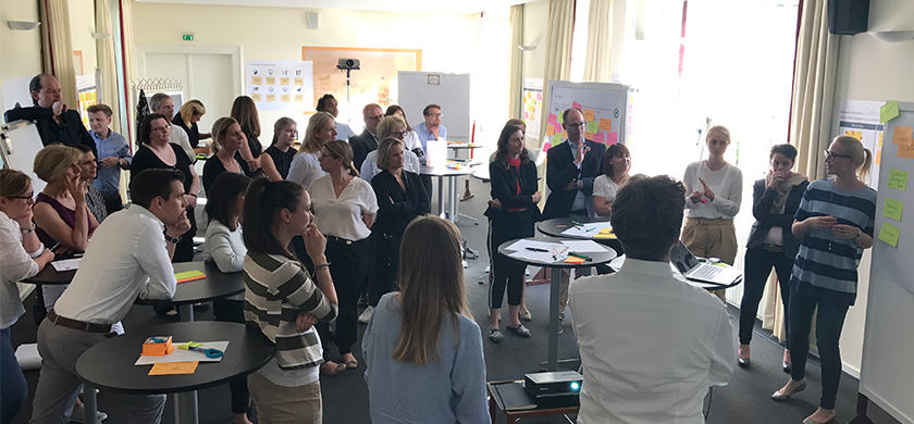 Eine Gruppe von Menschen bei einer Besprechung oder Workshop. Mehrere Teilnehmer stehen in einem Raum und hören einer Präsentation zu. Eine Frau erklärt etwas vor einer Wand mit bunten Post-it-Zetteln und Notizen. Einige Personen im Publikum stehen an Stehtischen und beobachten aufmerksam, während andere in Richtung der Präsentierenden schauen. Der Raum wirkt hell und modern, mit einem Projektor auf einem Tisch, der offenbar eine Präsentation anzeigt. Es ist eine interaktive Atmosphäre.
