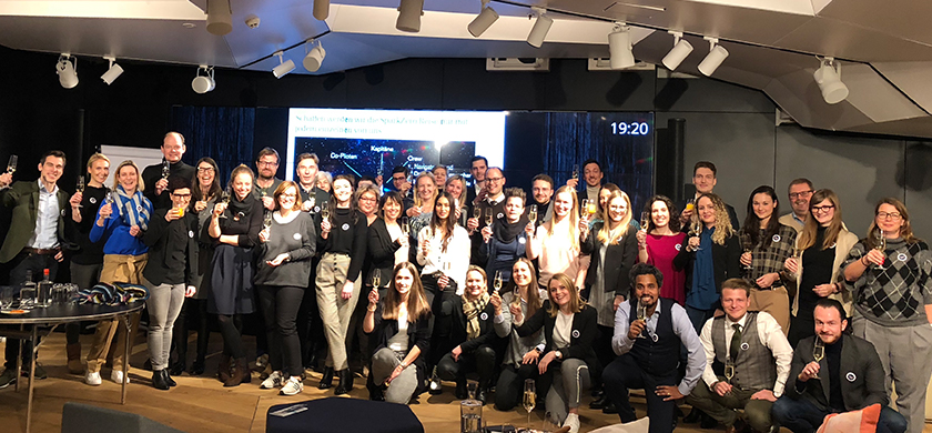 Eine Gruppe von BrandTrust Mitarbeiter, die zusammen auf einem Event oder Feier posieren. Die Mitarbeiter halten Gläser in der Hand und stoßen miteinander an. Es sind überwiegend junge Erwachsene in formeller oder halbformeller Kleidung zu sehen. Die Atmosphäre wirkt fröhlich und festlich. Im Hintergrund ist eine große Präsentationswand, was darauf hinweist, dass es sich um eine organisierte Veranstaltung handelt. Insgesamt entsteht ein freundlicher, gemeinschaftlicher Eindruck.