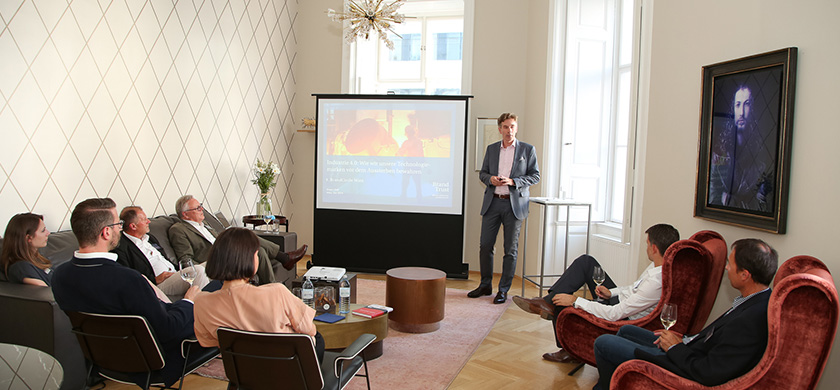Eine Präsentation in einem modernen, eleganten Raum. Ein Mann steht neben einem Projektor und erklärt etwas vor einer Gruppe von sieben Zuhörern, die auf einem Sofa und in Stühlen sitzen. Auf dem Bildschirm wird die Präsentation 'Industrie 4.0 und wie unsere Technologien den Arbeitsalltag verändern' angezeigt. Der Raum hat große Fenster, Holzboden und eine kunstvolle Wanddekoration. Der Präsentierende trägt einen Anzug und hält ein Glas in der Hand, während die Zuhörer aufmerksam zuhören.