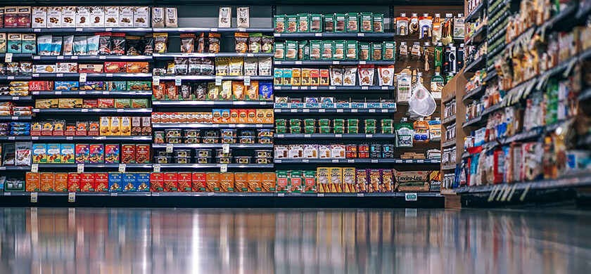 Supermarktregal mit verschiedenen Packungen von Snacks, Süßigkeiten, Keksen und anderen Lebensmitteln. Die Regale sind gut strukturiert, mit farbigen Verpackungen und Preisschildern, die deutlich sichtbar sind. Der Boden ist reflektierend, was eine klare Spiegelung des Regals zeigt.