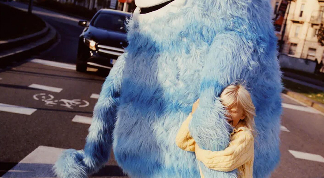 A child hugs a big blue monster and walks across the street with it.