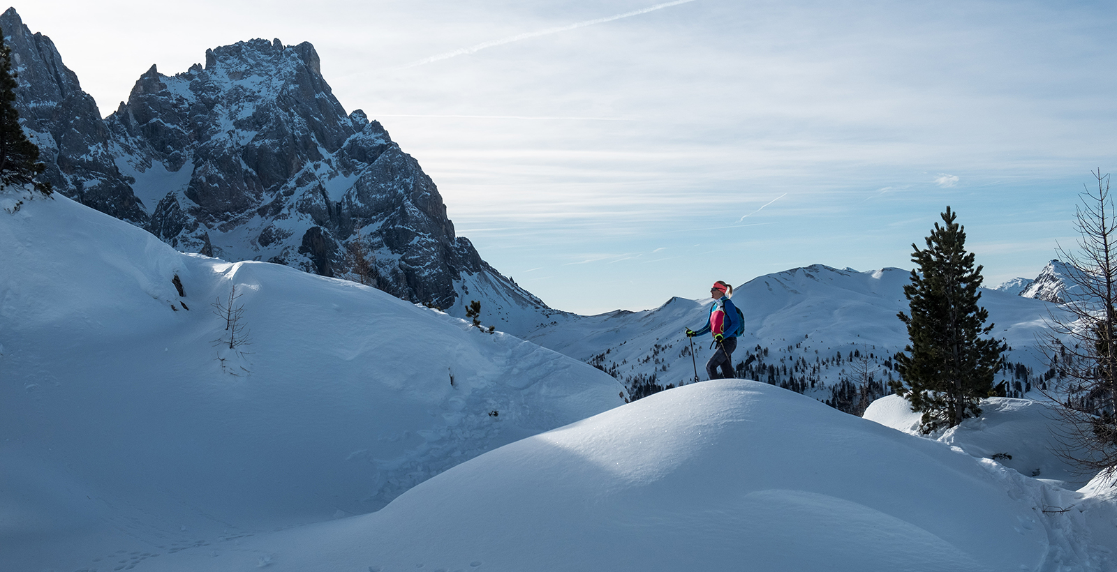 Person mit Skistöcken auf einer verschneiten Berglandschaft, unterwegs auf einem schmalen Pfad vor alpiner Bergkulisse.